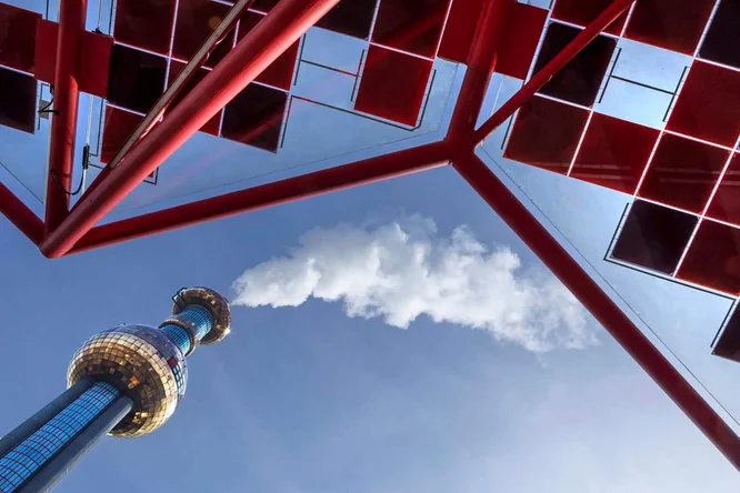 Ein hoher, spiegelnder Turm ragt in den klaren blauen Himmel. Von unten betrachtet, bildet er einen markanten roten Rahmen. Weiße Wolken oder Dampf steigen von der Turmspitze auf und erzeugen einen dynamischen visuellen Effekt. Die Szene besticht durch klare architektonische Linien und eine moderne, urbane Atmosphäre mit einer kühlen Farbpalette, akzentuiert durch ein leuchtendes Rot.