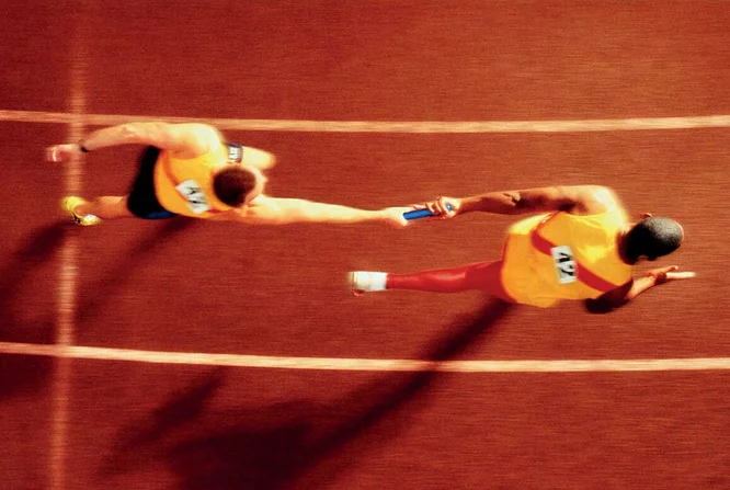 Overhead view of athletes performing a baton handoff during a relay race. The image captures the precision and teamwork required in track and field events, highlighting the dynamic movement and coordination between the runners.