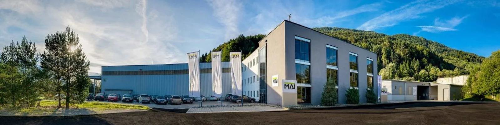 irmengebäude der Firma MAI mit geparkten Autos und Fahnen im Vordergrund an einem sonnigen Tag.