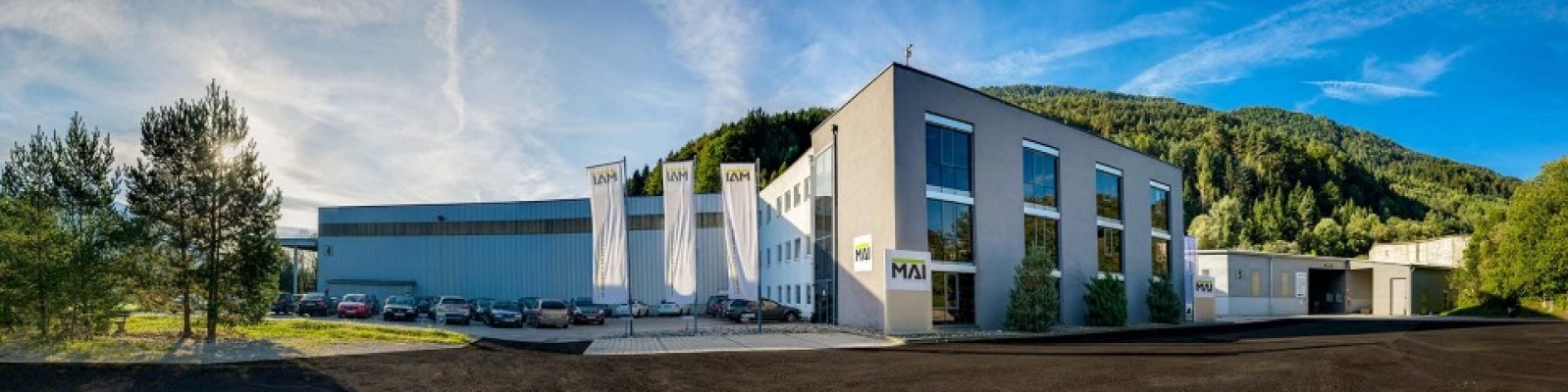 irmengebäude der Firma MAI mit geparkten Autos und Fahnen im Vordergrund an einem sonnigen Tag.