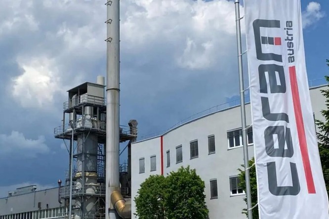 Das Bild zeigt eine Industrieanlage mit einem markanten Schornstein und einem angrenzenden modernen Gebäude. Eine große, vertikale Fahne im Vordergrund trägt die Aufschrift „Ceram Austria“ in fetten Buchstaben. Der Himmel ist leicht bewölkt, und grüne Bäume in der Nähe des Gebäudes bilden einen Kontrast zum industriellen Umfeld.