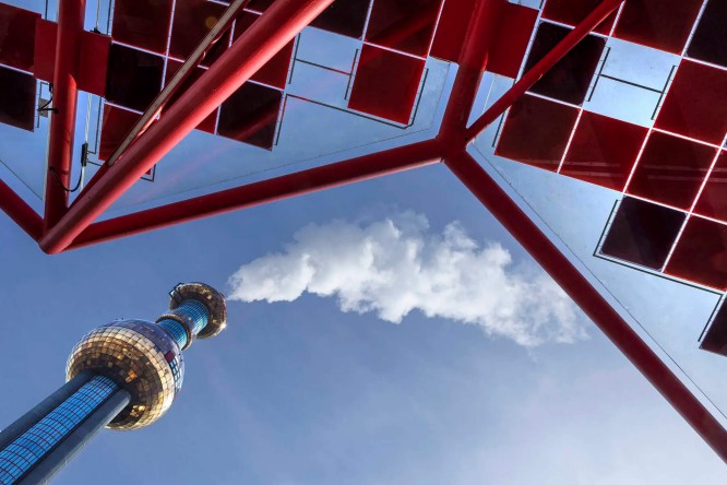 Ein hoher, spiegelnder Turm ragt in den klaren blauen Himmel. Von unten betrachtet, bildet er einen markanten roten Rahmen. Weiße Wolken oder Dampf steigen von der Turmspitze auf und erzeugen einen dynamischen visuellen Effekt. Die Szene besticht durch klare architektonische Linien und eine moderne, urbane Atmosphäre mit einer kühlen Farbpalette, akzentuiert durch ein leuchtendes Rot.